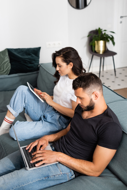 bearded freelancer using laptop near girlfriend with digital tablet in living room  - Foto, Imagem