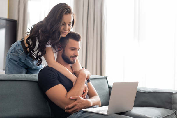 happy girl hugging handsome boyfriend using laptop in living room  - Foto, imagen