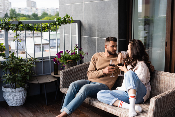 homem bonito e mulher atraente sentado no sofá ao ar livre e segurando copos de vinho enquanto olha um para o outro
 - Foto, Imagem