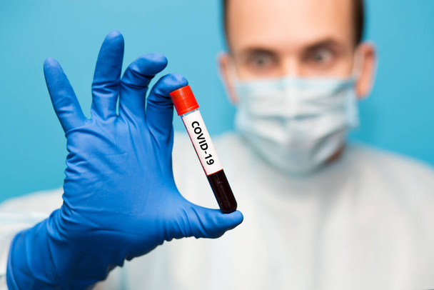 Un médecin portant un masque protecteur et des gants isolés sur fond bleu tient un tube à essai contenant un échantillon de sang ayant subi un test de dépistage du coronavirus. Tests de laboratoire sang des patients pour détecter la présence du coronavirus COVID-19
. - Photo, image