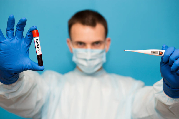Arzt in Schutzmaske und Handschuhen hält medizinisches Thermometer-Set 40 und Reagenzglas mit Blut auf Coronavirus isoliert auf blau getestet. Laboruntersuchung von Blut von Patienten auf das Vorhandensein von Coronavirus COVID-19. - Foto, Bild