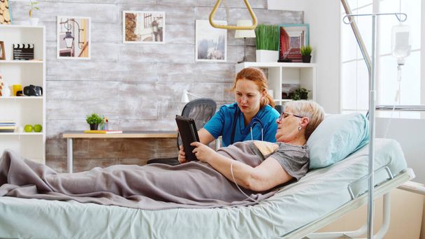 Opiekunka pomaga starszej niepełnosprawnej kobiecie leżącej w łóżku szpitalnym korzystać z tabletu cyfrowego Pc - Zdjęcie, obraz