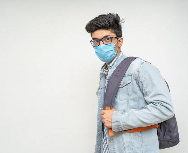 若いハンサムなアジアの少年は彼の大学の準備ができて、彼の背中にマスク、バッグを着て、カメラを見て. - 写真・画像
