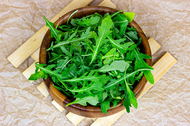 Top view of fresh green spicy arugula salad leaves in brown wooden bowl on light background. - Fotó, kép