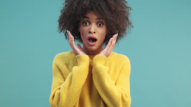 A surprised young african american woman with curly hair is opening her mouth and looking to the camera isolated over blue wall background in studio - Filmati, video