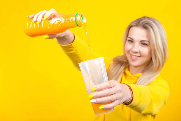 Joyeux belle jeune femme blonde en jaune casual vêtements sportifs versant du jus d'orange de bouteille en plastique au verre, fille souriante isolée sur fond jaune
. - Photo, image