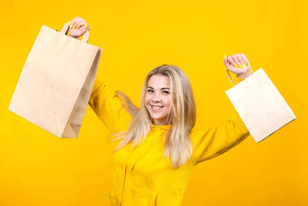 Portrait of young happy caucasian blonde woman with paper eco bags in yellow sportive suit, isolated on yellow background, smiling, looking at camera. - Foto, Bild