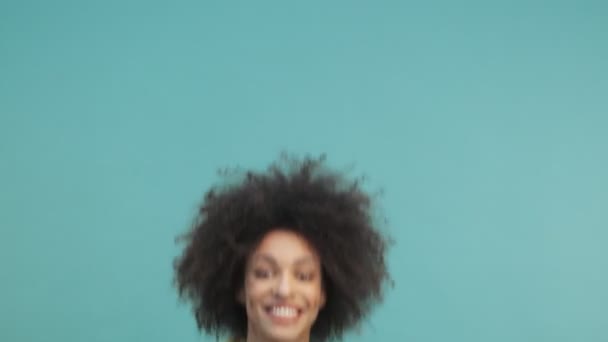 A cheerful young african american woman with curly hair is jumping and rejoicing isolated over blue wall background in studio - Metraje, vídeo