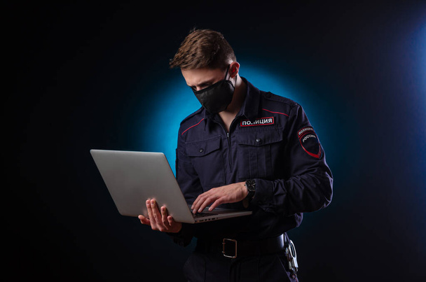 Ένας όμορφος άντρας με αστυνομική στολή με ένα λάπτοπ στο πρόσωπό του και μια ιατρική μάσκα. Αγγλική μετάφραση Police  - Φωτογραφία, εικόνα