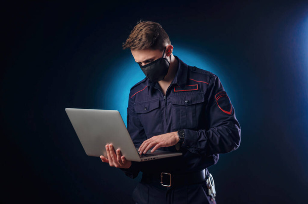 un tipo guapo en uniforme de policía con un portátil en la cara y una máscara médica
 . - Foto, imagen
