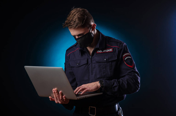 Een knappe man in een politie-uniform met een laptop op zijn gezicht en een medisch masker. Engelse vertaling van Police  - Foto, afbeelding
