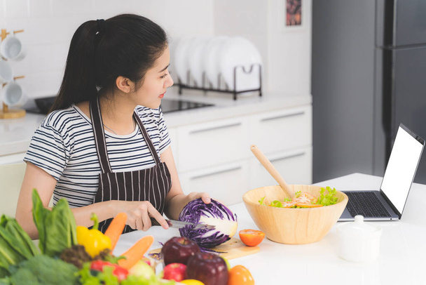Beautiful young woman is learning to cook healthy food online by the internet from a laptop in white kitchen with many fresh vegetables and fruits on table. - Photo, Image