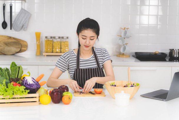Hermosa mujer joven está aprendiendo a cocinar alimentos saludables en línea por Internet desde un ordenador portátil en la cocina blanca con muchas verduras frescas y frutas en la mesa
. - Foto, imagen