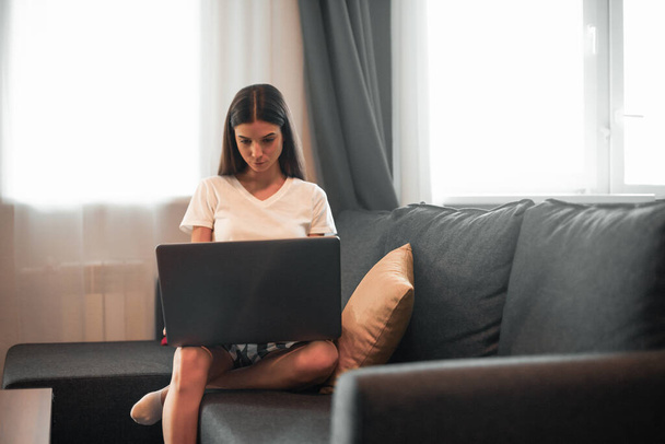 Young woman with modern laptop sitting on sofa at home. Happy girl browsing through the internet during free time..  - Fotó, kép