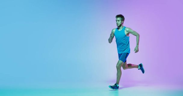 Full length portrait of active young caucasian running, jogging man on gradient studio background in neon light - Фото, зображення