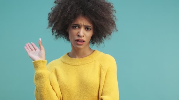 A bored emotional young african american woman with curly hair is gesturing her hand isolated over blue wall background in studio. Showing nonsense, bored, disgusting content - Materiał filmowy, wideo