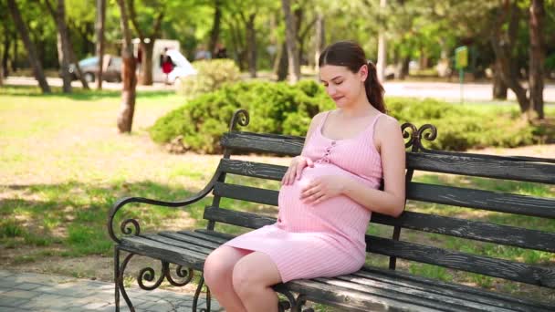 Κοντινό πλάνο των εγκύων γυναικών χέρια χάιδεμα κοιλιά σε ένα παγκάκι στο πάρκο το καλοκαίρι - Πλάνα, βίντεο