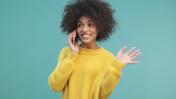 A smiling positive young african american woman with curly hair is talking on the phone isolated over blue wall background in studio - Séquence, vidéo