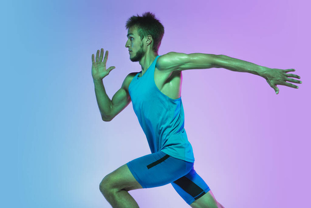 Full length portrait of active young caucasian running, jogging man on gradient studio background in neon light - Photo, Image