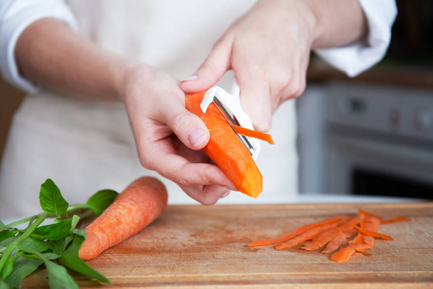 Hands of a young woman in an apron, kitchen background, peeling vegetables or carrot using a food peeler. Cooking before serving. - Foto, Bild