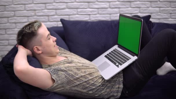 Happy Young Man ontspannen op de bank, op zoek laptop groen scherm mockup, gezellig huis - Video