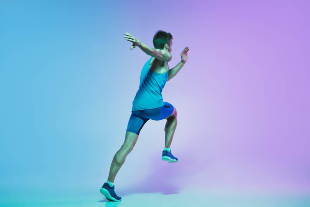 Full length portrait of active young caucasian running, jogging man on gradient studio background in neon light - 写真・画像