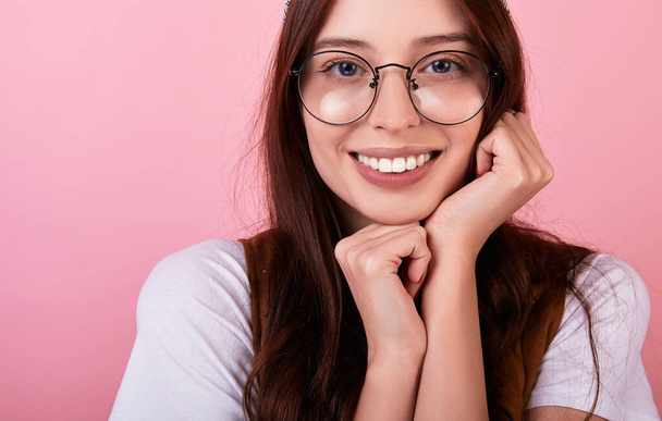 Portrait of a beautiful long-haired brunette hipster student in a bandana, glasses, vest on a pink background. Lady emotionally presses her hands to her face,feeling safe and care close-up.Copy space. - Photo, image