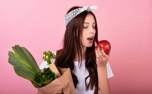 Cute young Caucasian girl vegan hipster, enjoys eating fresh tasty red apple and holds green vegetables from an eco farm, lifestyle, diet in joy, healthy food. In a basic T-shirt on a pink background. - Foto, immagini