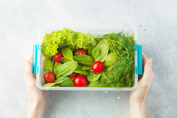 Diverse groente voor salade - sla, spinazie, dille en kerstomaten in groentekistje met vrouwelijke handen op grijze stenen ondergrond. Voedselbereiding. Groen eten concept. Bovenaanzicht - Foto, afbeelding