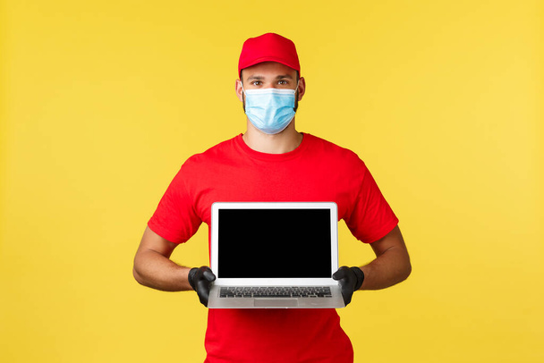 Express delivery during pandemic, covid-19, safe shipping, online shopping concept. Employee in red uniform cap and gloves, courier in medical mask showing website on laptop screen - Foto, imagen