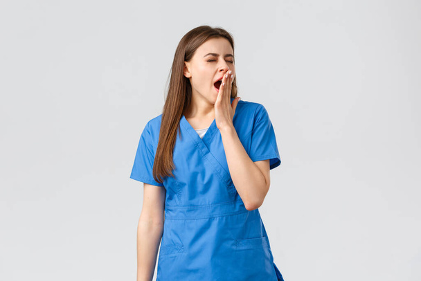 Healthcare workers, prevent virus, insurance and medicine concept. Tired young female nurse had tough night shift at hospital, yawning, cover opened mouth with palm, feel sleepy, lack energy - Photo, Image