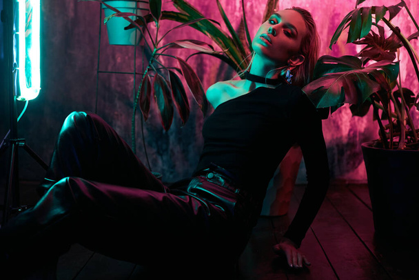 Fashion woman sitting on the floor in tropical foliage neon light. Wet hair, perfect figure and makeup. Pink and green neon color. Beautiful girl and monstera leaves - Foto, imagen