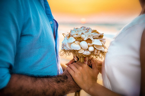 Buquê de casamento (feito de conchas) nas mãos da noiva e do noivo, pôr do sol do mar no fundo
. - Foto, Imagem