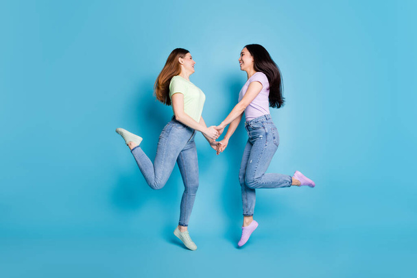 Full body profile photo of two people lesbians couple ladies jump high hold arms look eyes good mood happy together wear casual t-shirts jeans footwear isolated blue color background - Fotoğraf, Görsel