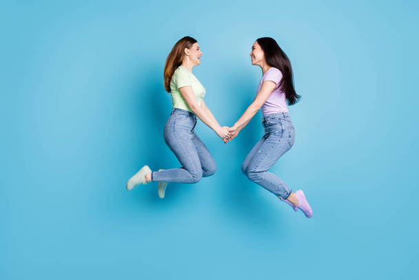Full body profile photo of two people lesbians couple ladies jump high up hold hands good mood rejoicing happy together wear casual t-shirts jeans footwear isolated blue color background - Fotografie, Obrázek