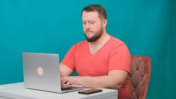 O homem de negócios feliz jovem em uma camiseta rosa trabalha em um computador portátil em uma tela verde. Retrato de um homem que fala a olhar para o portátil. Homem trabalhando em sua mesa no escritório
. - Foto, Imagem