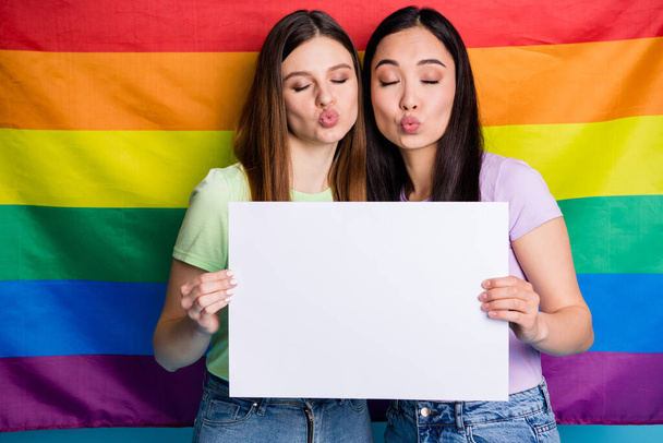 Photo of charming flirty lesbians couple ladies protest parade day tolerance same sex marriage send air kiss gay rainbow flag background hold billboards protesters wear casual t-shirts jeans - Фото, изображение