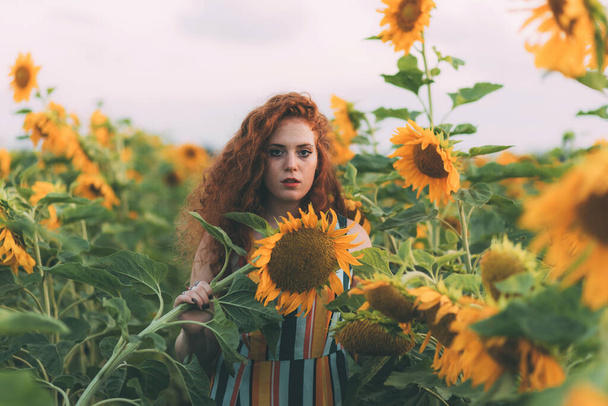 Beautiful young girl with red wavy hair and freckles in stripped colourful dress enjoying nature on the field of sunflowers. The concept of summer and sun. Sunflower season. Holding sunflower - Fotó, kép