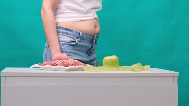 worried woman with a fat belly should choose between a green apple and a sausage. Dieting concept, beautiful young woman choosing between healthy food and junk food - Footage, Video