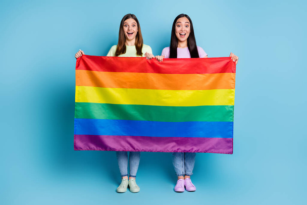 Full size photo of funny cheerful lesbians couple came foreign parade support tolerance same sex marriages hold gay rainbow flag wear casual t-shirts jeans isolated blue color background - Valokuva, kuva