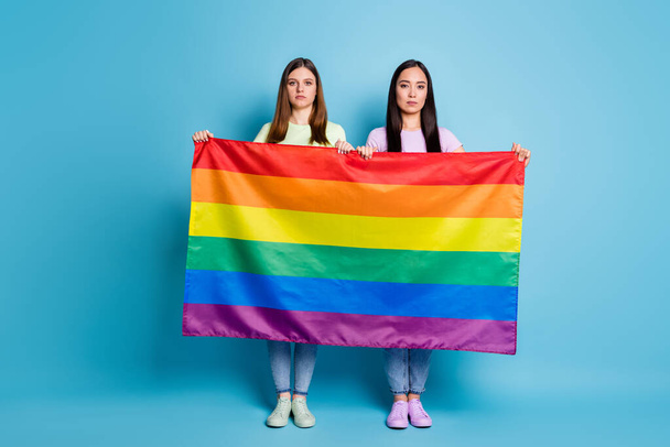 Full size photo of serious free love lesbians couple ladies came foreign parade tolerance same sex marriages hold gay rainbow flag wear casual t-shirts jeans isolated blue color background - Foto, Imagem