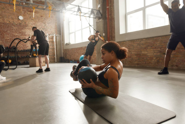 Fitness is my Outlook. Athletic young woman using a ball while having workout on a mat at industrial gym. Group training, teamwork concept - Photo, image