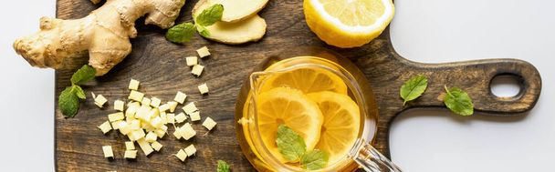top view of hot tea near ginger root, lemon and mint on wooden cutting board on white background, panoramic shot - Photo, Image