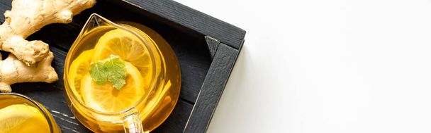 vrchní pohled na horký čaj s kořenem zázvoru, citron a máta na dřevěném podnosu na bílém pozadí, panoramatický záběr - Fotografie, Obrázek