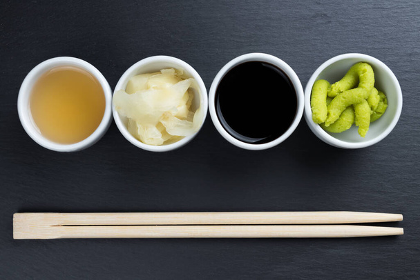Τζίντζερ, ιαπωνικό horseradish wasabi, σόγια και σάλτσα ψαριού σε μικρά λευκά μπολ με chopsticks σε μαύρο πιάτο σχιστόλιθου κουζίνας. Αντιγραφή χώρου στο κάτω μέρος. Ασιατική κουζίνα αφηρημένη έννοια. - Φωτογραφία, εικόνα