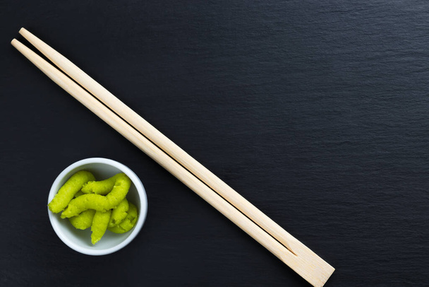 Japanse mierikswortel wasabi in kleine witte kom met eetstokjes op zwarte keukenblad. Typische Aziatische keuken kruiden. Kopieer ruimte in de rechterbovenhoek. - Foto, afbeelding