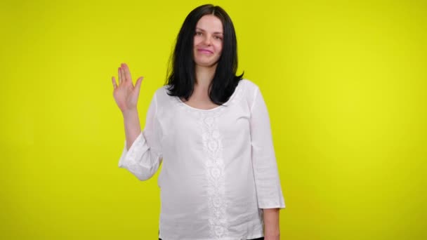 Hallo oder Tschüss. Junge schwangere Frau lächelt und schüttelt Hand auf gelbem Hintergrund - Filmmaterial, Video