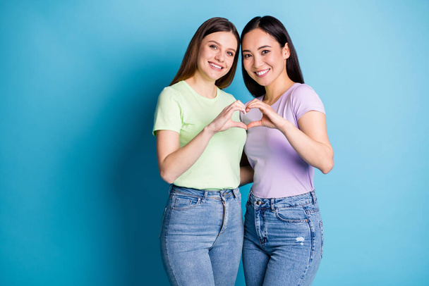 Photo of affectionate lesbians couple young ladies romantic date make arms fingers heart shape symbol lovers anniversary wear casual t-shirts jean isolated blue color background - Foto, Imagen