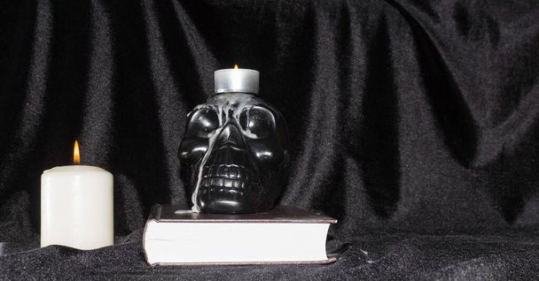 El concepto de magia, ocultismo y esoterismo. Un cráneo humano negro en un libro con velas sobre un fondo negro, espacio para copiar
. - Foto, imagen
