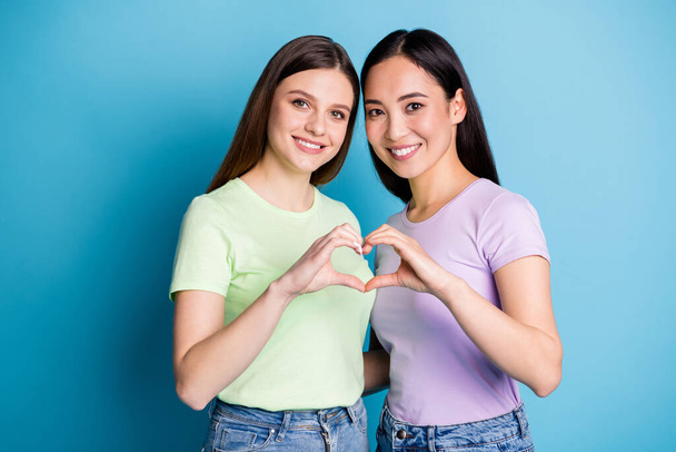 Photo of two affectionate lesbians couple young students ladies romance date making arms fingers heart shape lovers anniversary wear casual t-shirts isolated blue color background - Foto, immagini